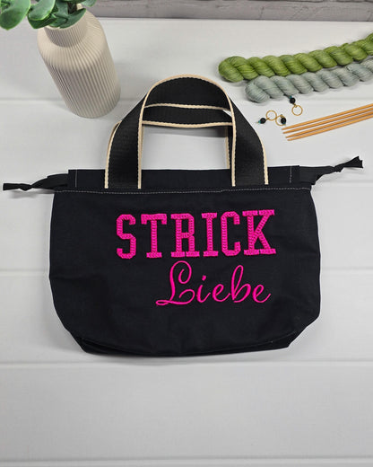 Projekttasche Alva S aus schwarzem Canvas mit erhabenem 3D-Stickzug Strickliebe in Pink, Henkeln und Kordelzugkragen. Handgenäht in Bayern von Nadelmikado.
