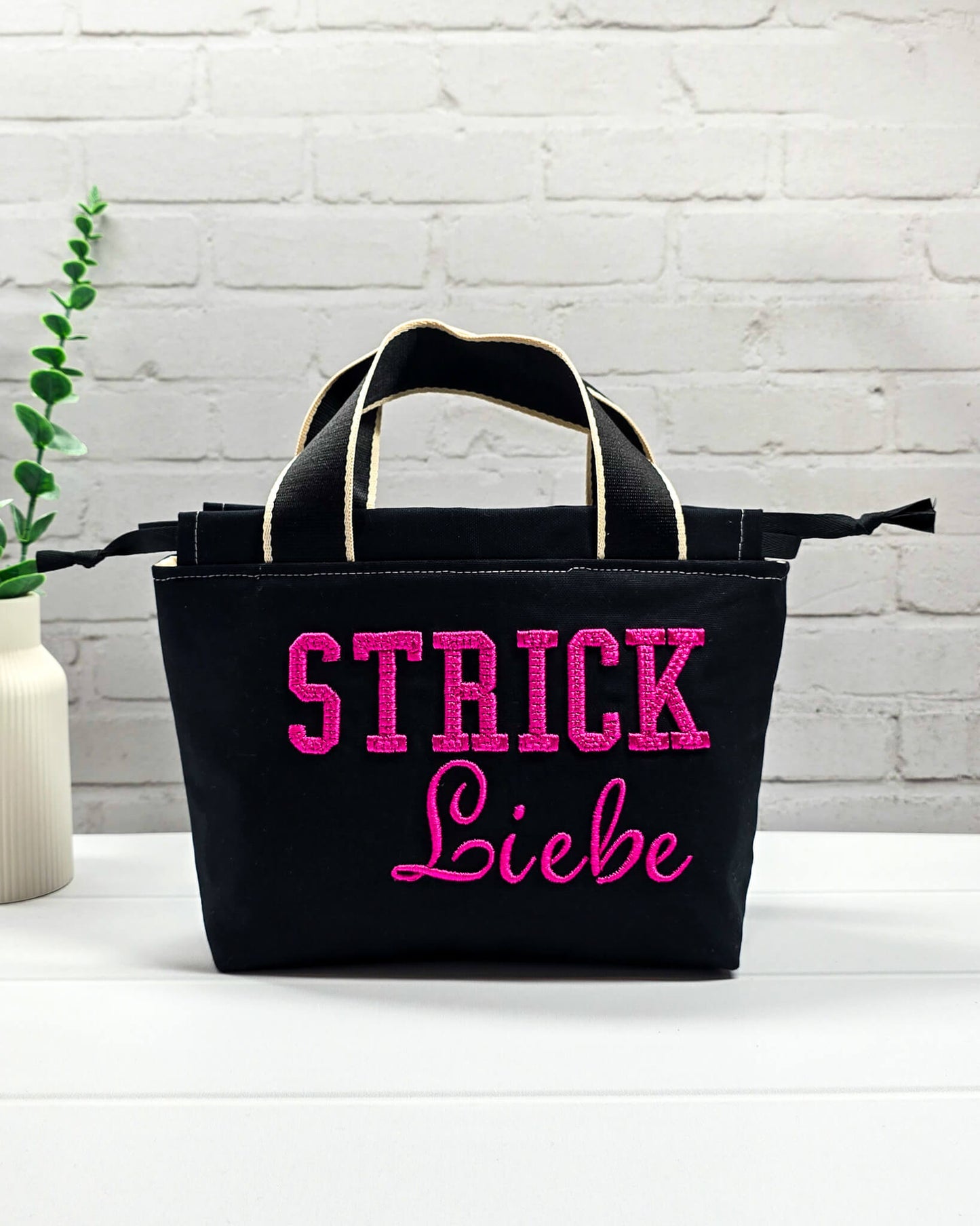 Projekttasche Alva S aus schwarzem Canvas mit erhabenem 3D-Stickzug Strickliebe in Pink, Henkeln und Kordelzugkragen. Handgenäht in Bayern von Nadelmikado.