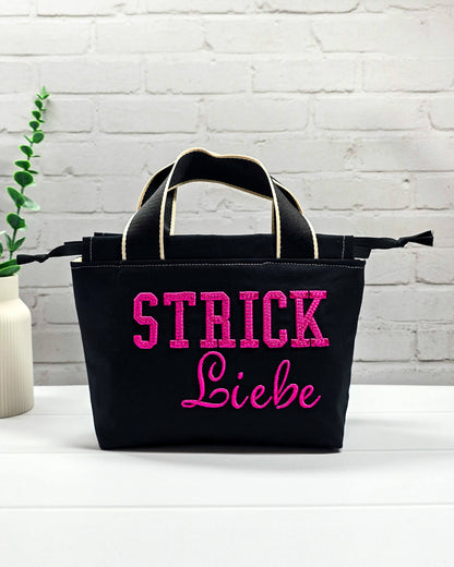 Projekttasche Alva S aus schwarzem Canvas mit erhabenem 3D-Stickzug Strickliebe in Pink, Henkeln und Kordelzugkragen. Handgenäht in Bayern von Nadelmikado.