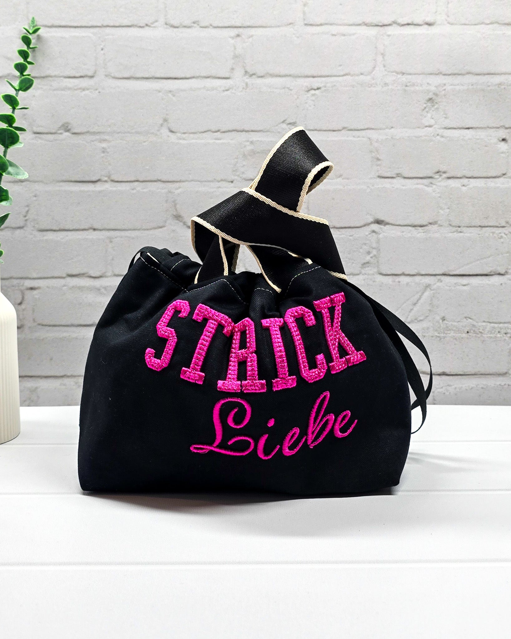Projekttasche Alva S aus schwarzem Canvas mit erhabenem 3D-Stickzug Strickliebe in Pink, Henkeln und Kordelzugkragen. Handgenäht in Bayern von Nadelmikado.