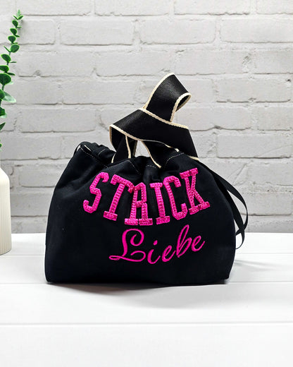 Projekttasche Alva S aus schwarzem Canvas mit erhabenem 3D-Stickzug Strickliebe in Pink, Henkeln und Kordelzugkragen. Handgenäht in Bayern von Nadelmikado.