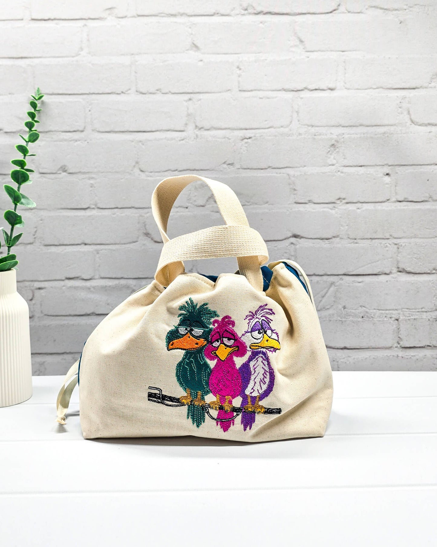 Projekttasche Alva S aus hellem Canvas mit Stickmotiv drei bunte Vögel auf Stange, Henkeln und petrolblauem Kordelzug. Handgenäht in Bayern von Nadelmikado.