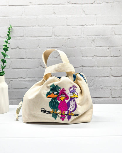 Projekttasche Alva S aus hellem Canvas mit Stickmotiv drei bunte Vögel auf Stange, Henkeln und petrolblauem Kordelzug. Handgenäht in Bayern von Nadelmikado.