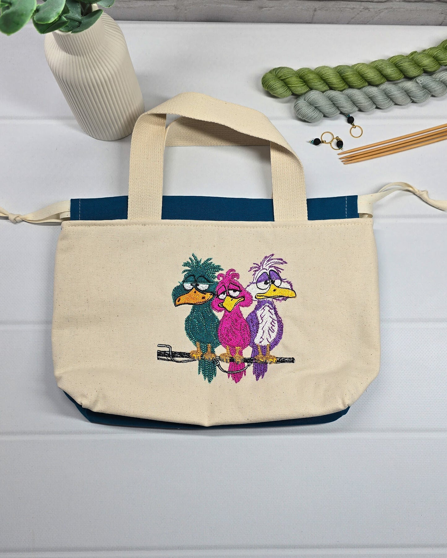 Projekttasche Alva S aus hellem Canvas mit Stickmotiv drei bunte Vögel auf Stange, Henkeln und petrolblauem Kordelzug. Handgenäht in Bayern von Nadelmikado.