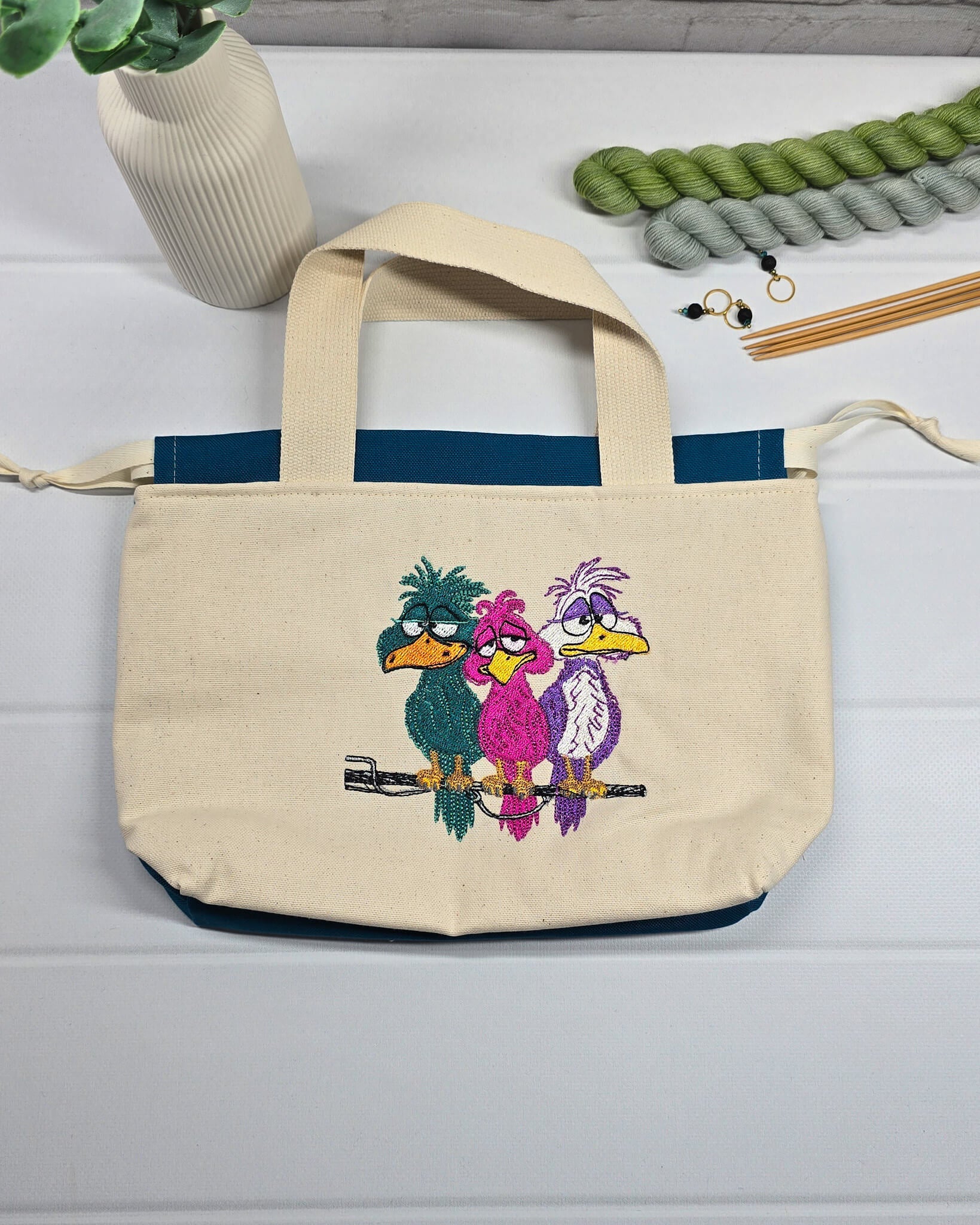 Projekttasche Alva S aus hellem Canvas mit Stickmotiv drei bunte Vögel auf Stange, Henkeln und petrolblauem Kordelzug. Handgenäht in Bayern von Nadelmikado.