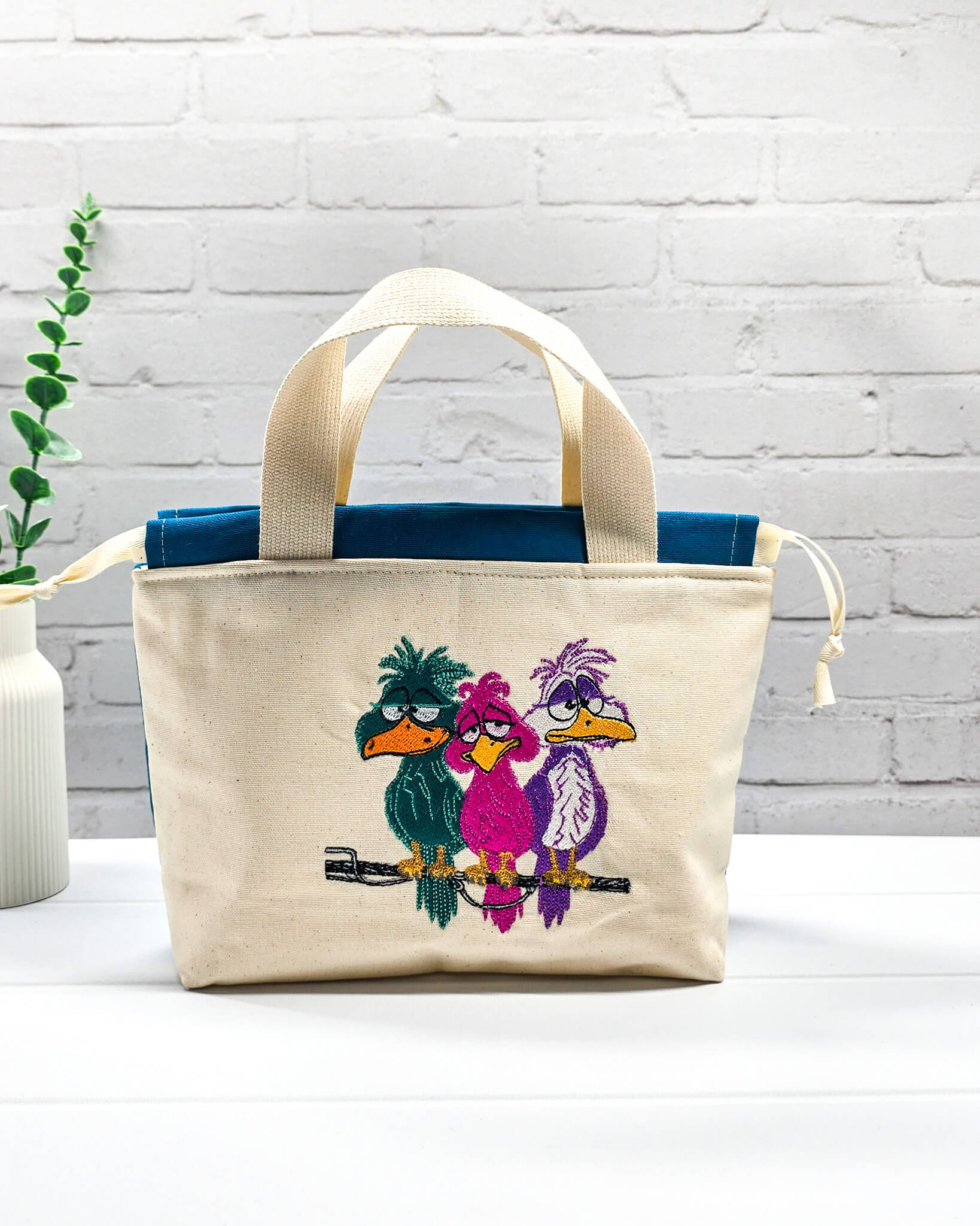 Projekttasche Alva S aus hellem Canvas mit Stickmotiv drei bunte Vögel auf Stange, Henkeln und petrolblauem Kordelzug. Handgenäht in Bayern von Nadelmikado.