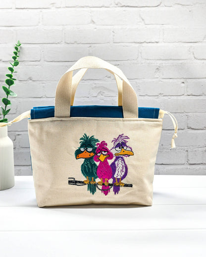 Projekttasche Alva S aus hellem Canvas mit Stickmotiv drei bunte Vögel auf Stange, Henkeln und petrolblauem Kordelzug. Handgenäht in Bayern von Nadelmikado.