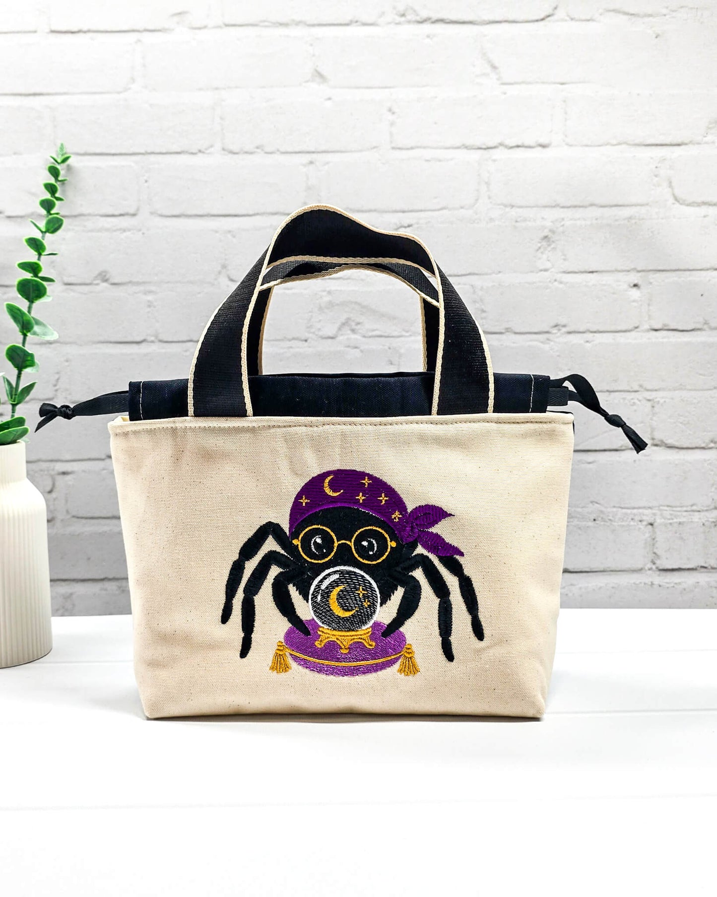 Projekttasche Alva S aus hellem Canvas mit gestickter Wahrsager Spinne und Kristallkugel, Henkeln und schwarzem Kordelzugkragen. Handgenäht in Bayern von Nadelmikado.