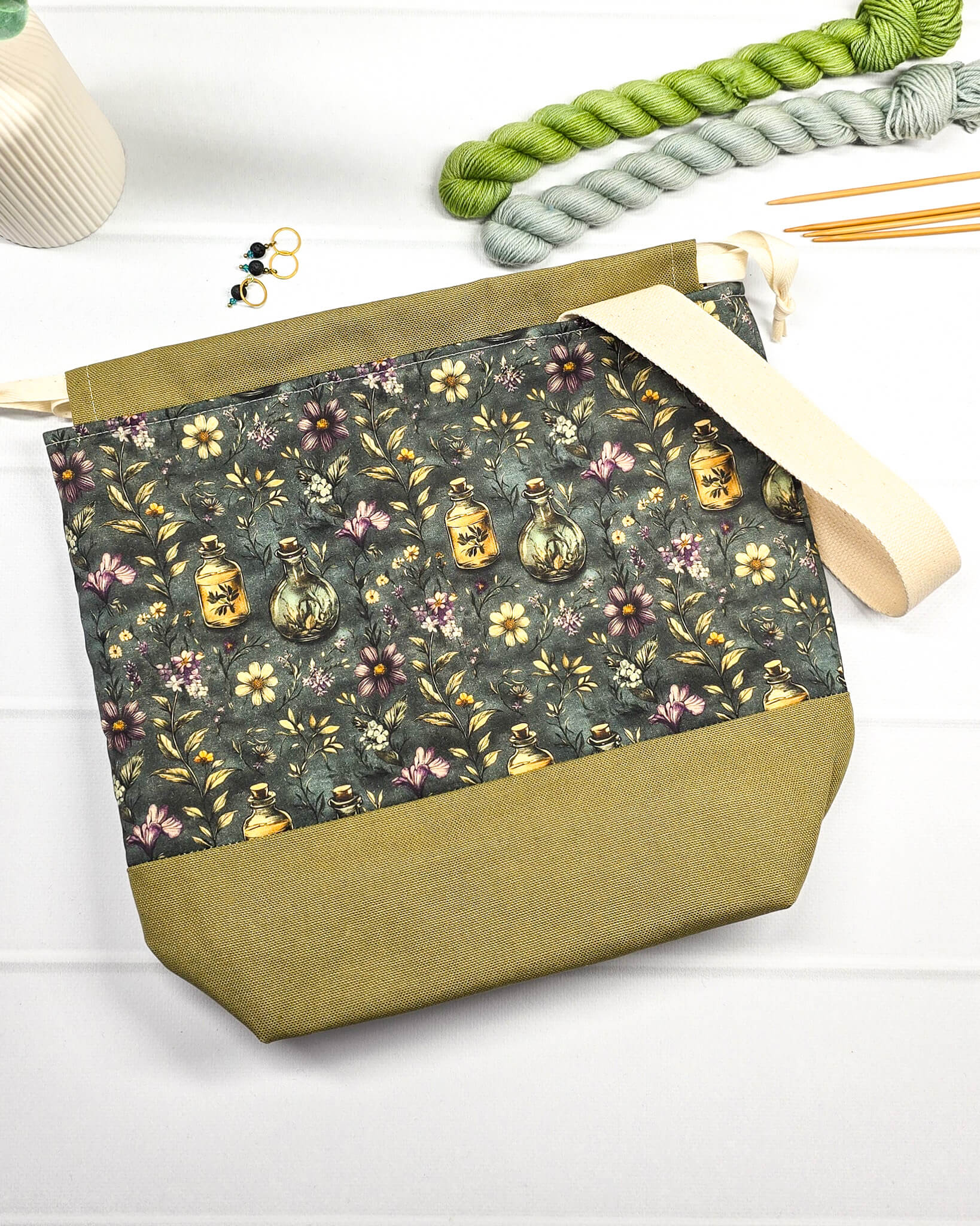 Projekttasche mit Kordelzug und cremefarbenem Tragegurt aus dunkelgrünem Stoff mit kleinen Blüten und Trankflaschen sowie olivgrünem Boden. Bietet Platz für 400g Wolle und schützt das Strickzeug vor Verheddern. Handgenäht in Bayern von Nadelmikado.