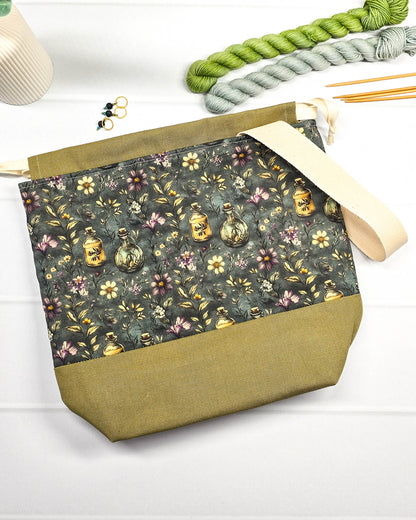 Projekttasche mit Kordelzug und cremefarbenem Tragegurt aus dunkelgrünem Stoff mit kleinen Blüten und Trankflaschen sowie olivgrünem Boden. Bietet Platz für 400g Wolle und schützt das Strickzeug vor Verheddern. Handgenäht in Bayern von Nadelmikado.