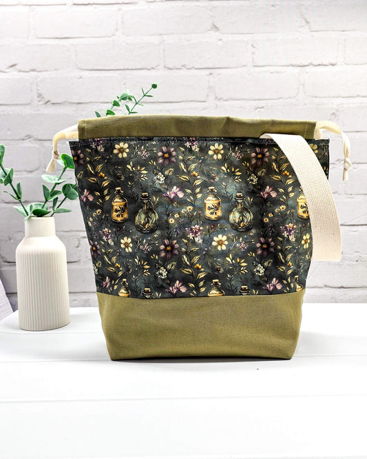 Projekttasche mit Kordelzug und cremefarbenem Tragegurt aus dunkelgrünem Stoff mit kleinen Blüten und Trankflaschen sowie olivgrünem Boden. Bietet Platz für 400g Wolle und schützt das Strickzeug vor Verheddern. Handgenäht in Bayern von Nadelmikado.