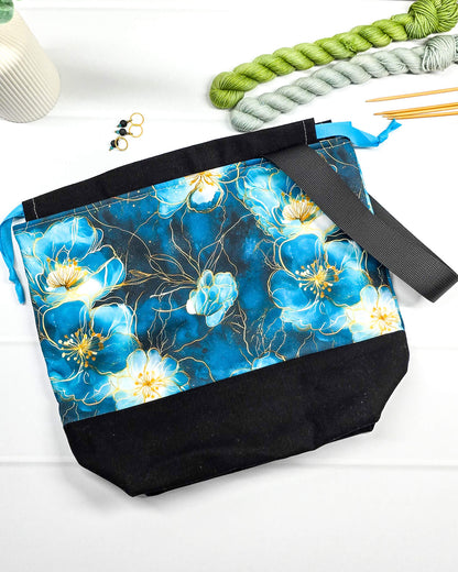 Projekttasche mit Kordelzug und schwarzem Tragegurt aus türkis-blauem Blumenstoff mit Goldakzenten und schwarzem Boden. Bietet Platz für 400g Wolle und schützt das Strickzeug vor Verschmutzung. Handgenäht in Bayern von Nadelmikado.