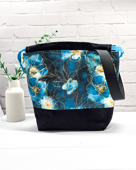 Projekttasche mit Kordelzug und schwarzem Tragegurt aus türkis-blauem Blumenstoff mit Goldakzenten und schwarzem Boden. Bietet Platz für 400g Wolle und schützt das Strickzeug vor Verschmutzung. Handgenäht in Bayern von Nadelmikado.