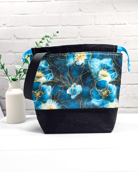 Projekttasche mit Kordelzug und schwarzem Tragegurt aus türkis-blauem Blumenstoff mit Goldakzenten und schwarzem Boden. Bietet Platz für 400g Wolle und schützt das Strickzeug vor Verschmutzung. Handgenäht in Bayern von Nadelmikado.