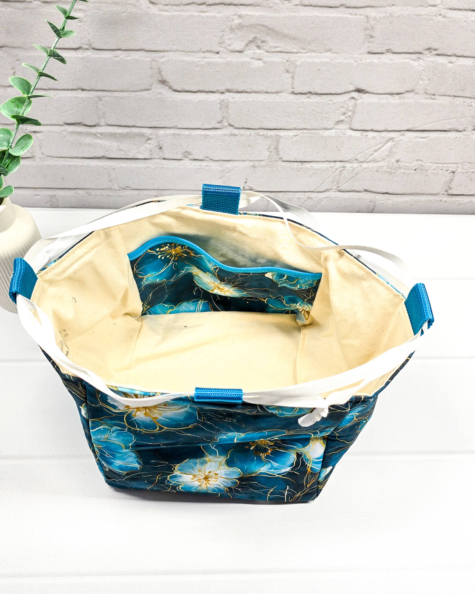 Projekttasche Aska M in Rice-Bag-Form mit großen blauen Blüten auf dunklem Stoff, hellen Verschlusskordeln und zwei blauen Tragegriffen. Handgenäht in Bayern von Nadelmikado. 