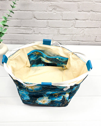 Projekttasche Aska M in Rice-Bag-Form mit großen blauen Blüten auf dunklem Stoff, hellen Verschlusskordeln und zwei blauen Tragegriffen. Handgenäht in Bayern von Nadelmikado. 