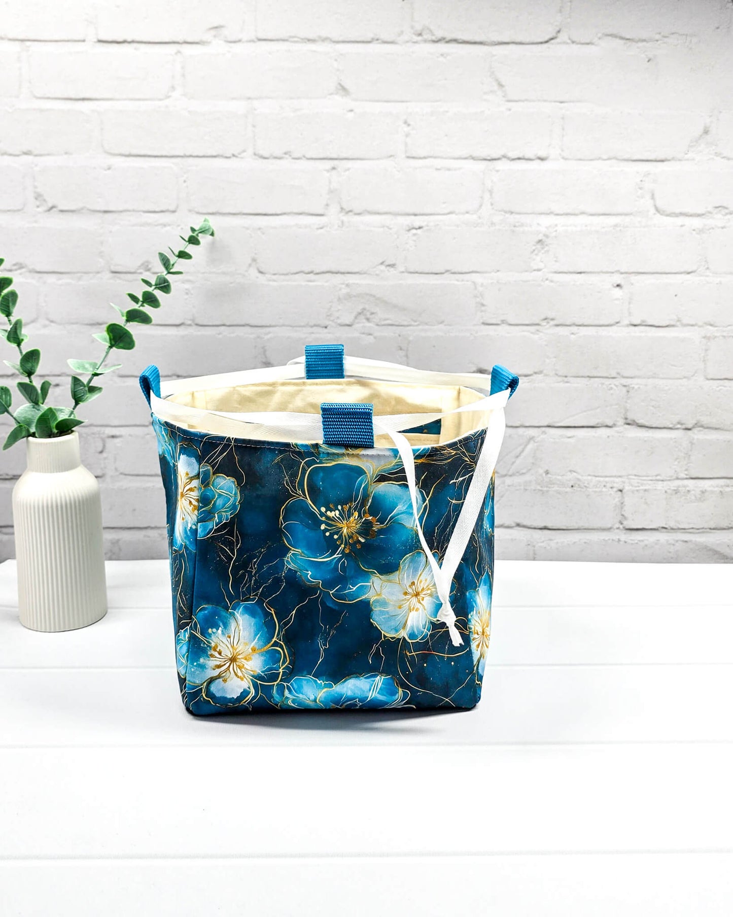 Projekttasche Aska M in Rice-Bag-Form mit großen blauen Blüten auf dunklem Stoff, hellen Verschlusskordeln und zwei blauen Tragegriffen. Handgenäht in Bayern von Nadelmikado. 