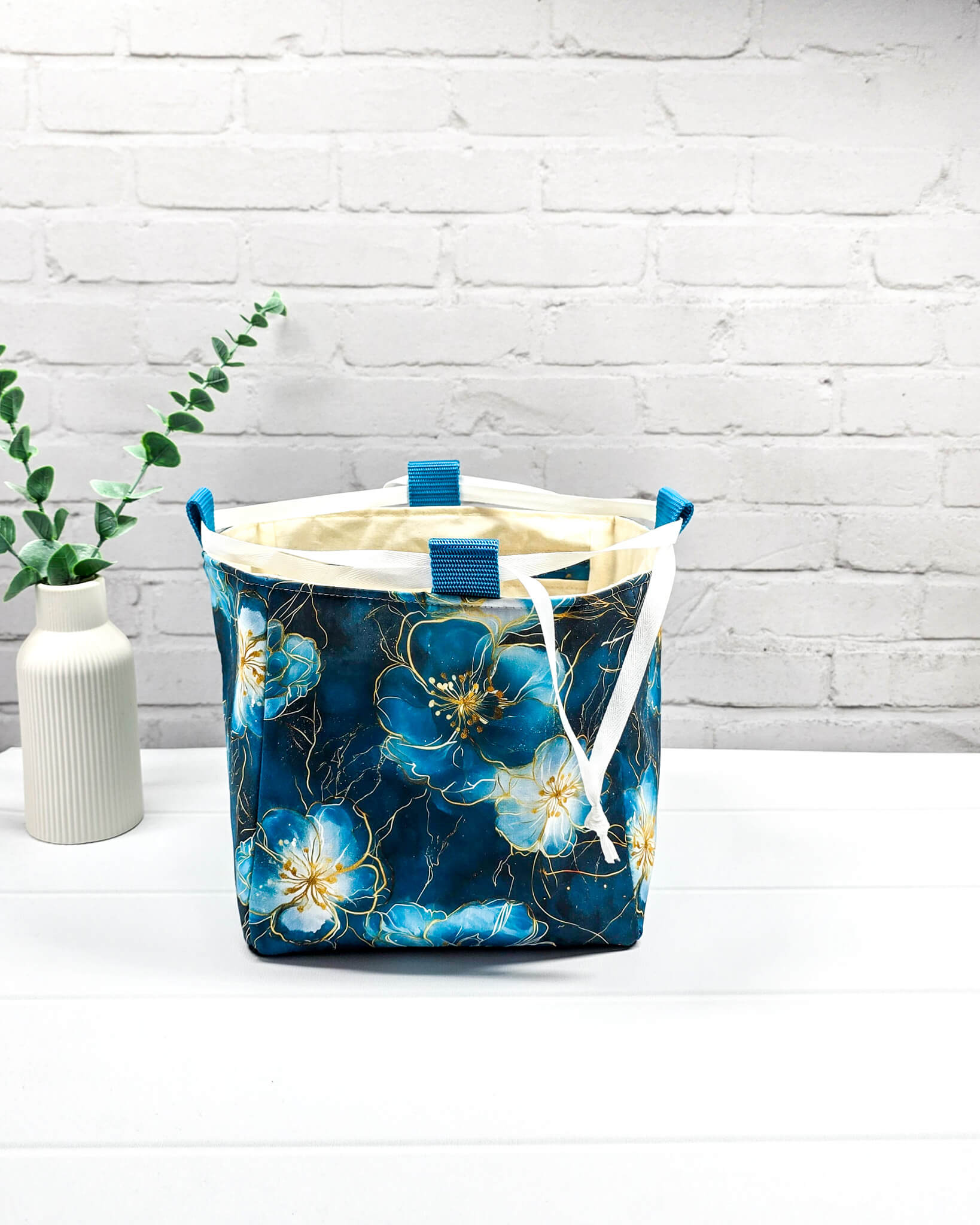 Projekttasche Aska M in Rice-Bag-Form mit großen blauen Blüten auf dunklem Stoff, hellen Verschlusskordeln und zwei blauen Tragegriffen. Handgenäht in Bayern von Nadelmikado. 