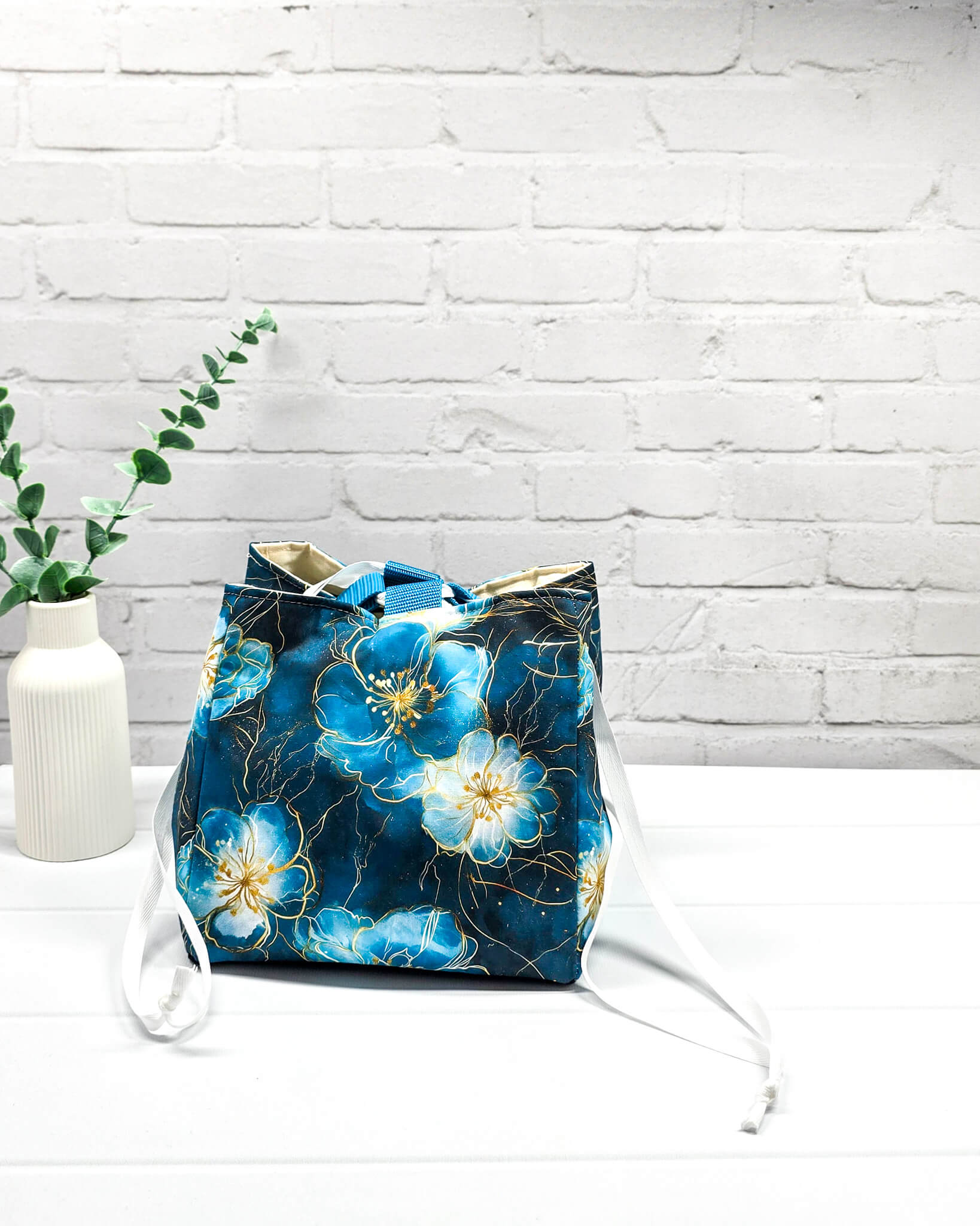 Projekttasche Aska M in Rice-Bag-Form mit großen blauen Blüten auf dunklem Stoff, hellen Verschlusskordeln und zwei blauen Tragegriffen. Handgenäht in Bayern von Nadelmikado. 