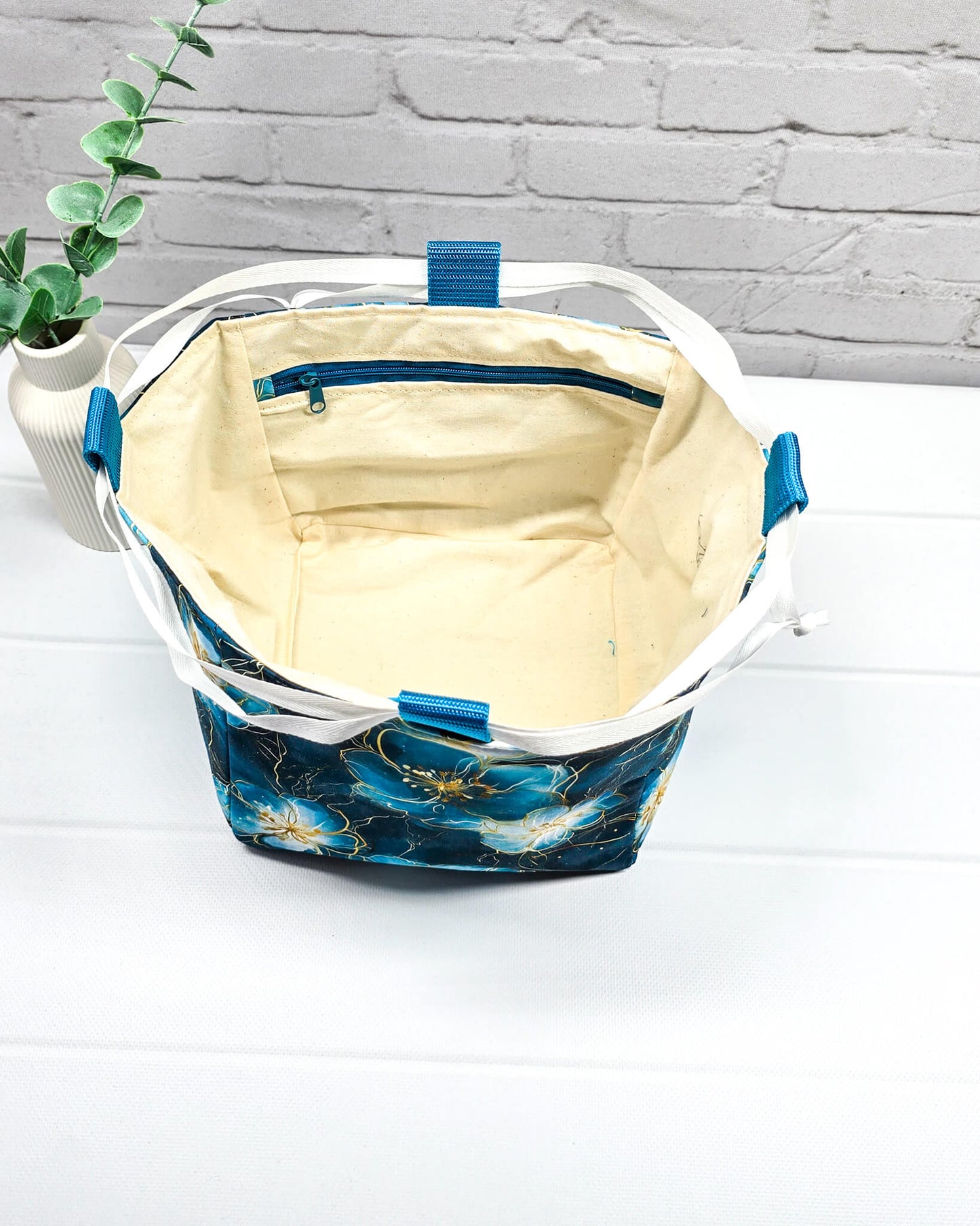 Projekttasche Aska M in Rice-Bag-Form mit großen blauen Blüten auf dunklem Stoff, hellen Verschlusskordeln und zwei blauen Tragegriffen. Handgenäht in Bayern von Nadelmikado. 