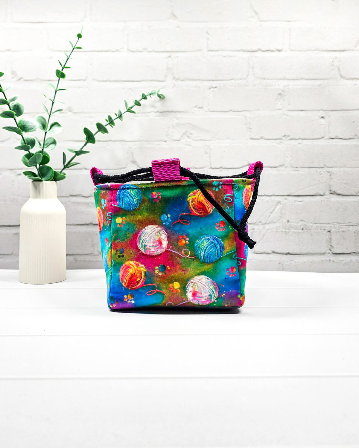 Projekttasche Aska Größe S als Rice Bag mit bunten Wollknäueln auf leuchtendem Stoff in Pink, Türkis, Blau und Grün. Mit schwarzem Kordelzug und pinker Schlaufe. Handgenäht in Bayern von Nadelmikado.