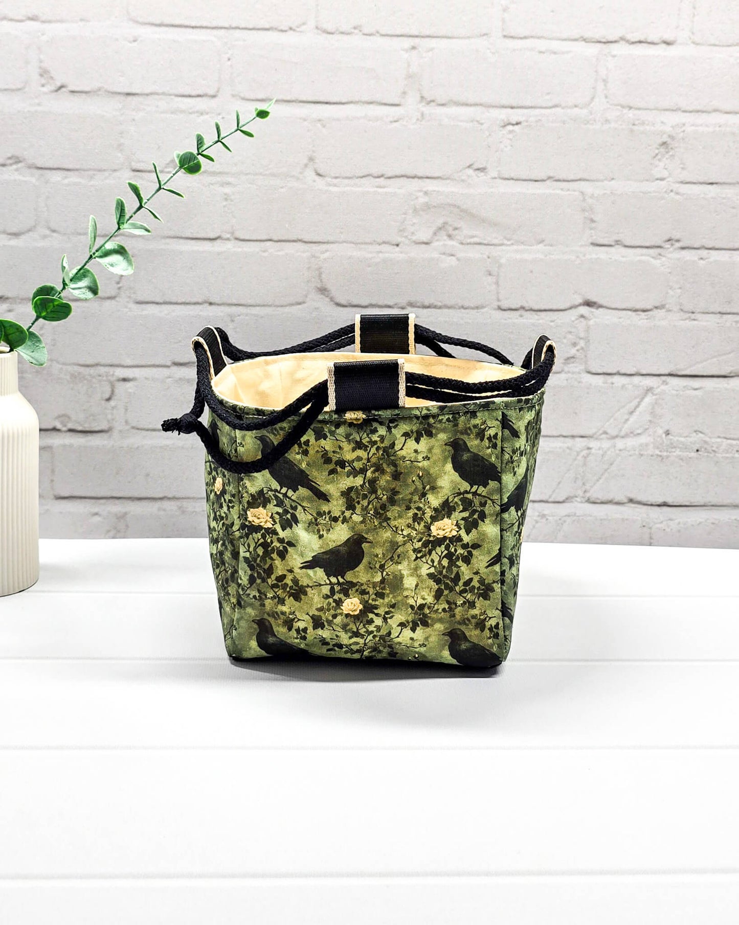 Kleine grüne Projekttasche mit Kordelzug im Rice Bag Stil, gemustert mit Raben und Rosen, genutzt als Strickbeutel für ein Sockenstrickprojekt mit Wolle und Nadelspiel. Handgenäht in Bayern von Nadelmikado.