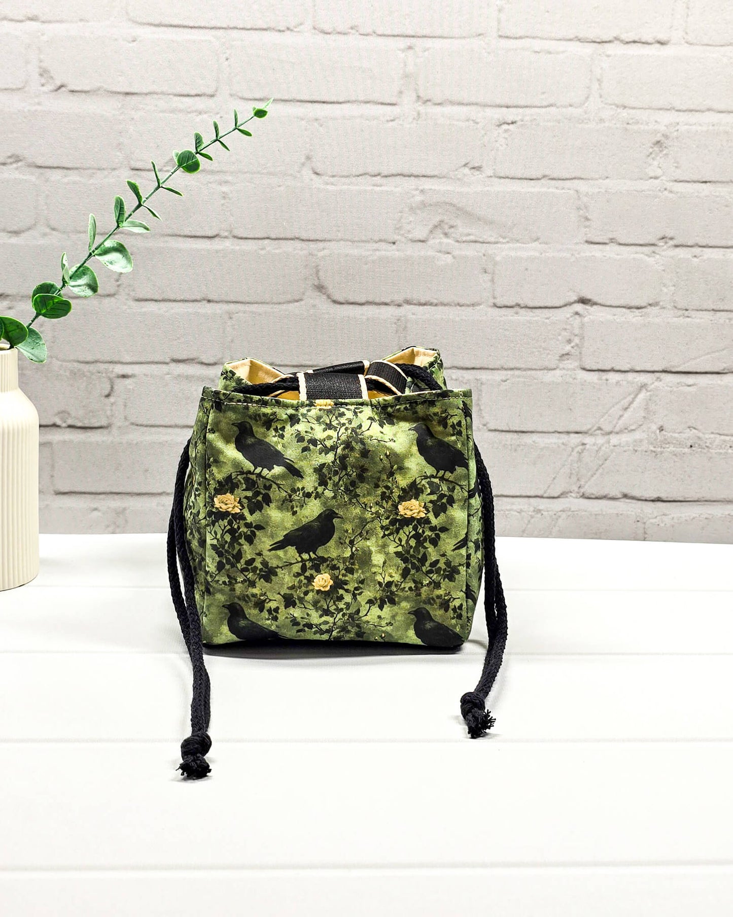 Kleine grüne Projekttasche mit Kordelzug im Rice Bag Stil, gemustert mit Raben und Rosen, genutzt als Strickbeutel für ein Sockenstrickprojekt mit Wolle und Nadelspiel. Handgenäht in Bayern von Nadelmikado.