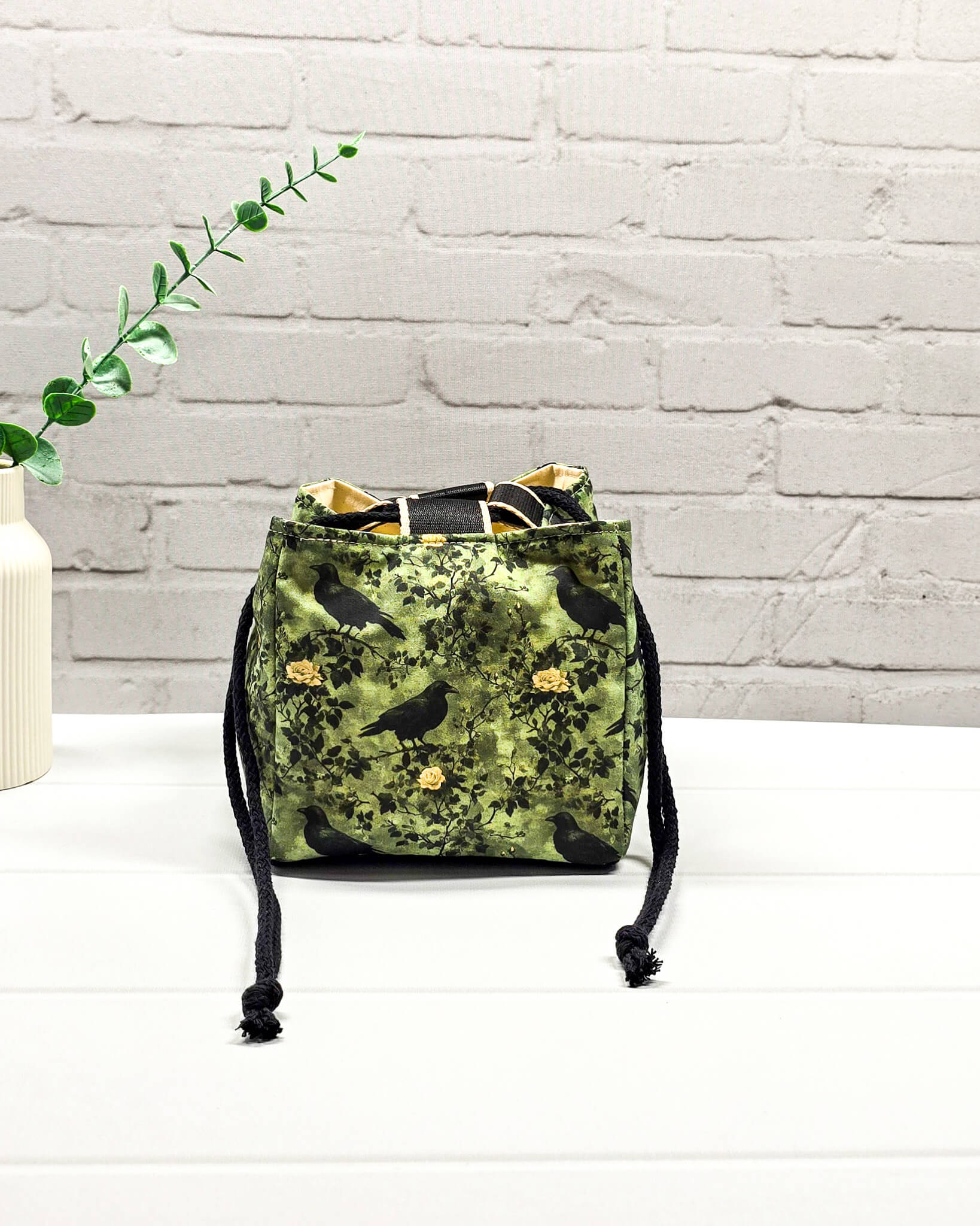 Kleine grüne Projekttasche mit Kordelzug im Rice Bag Stil, gemustert mit Raben und Rosen, genutzt als Strickbeutel für ein Sockenstrickprojekt mit Wolle und Nadelspiel. Handgenäht in Bayern von Nadelmikado.