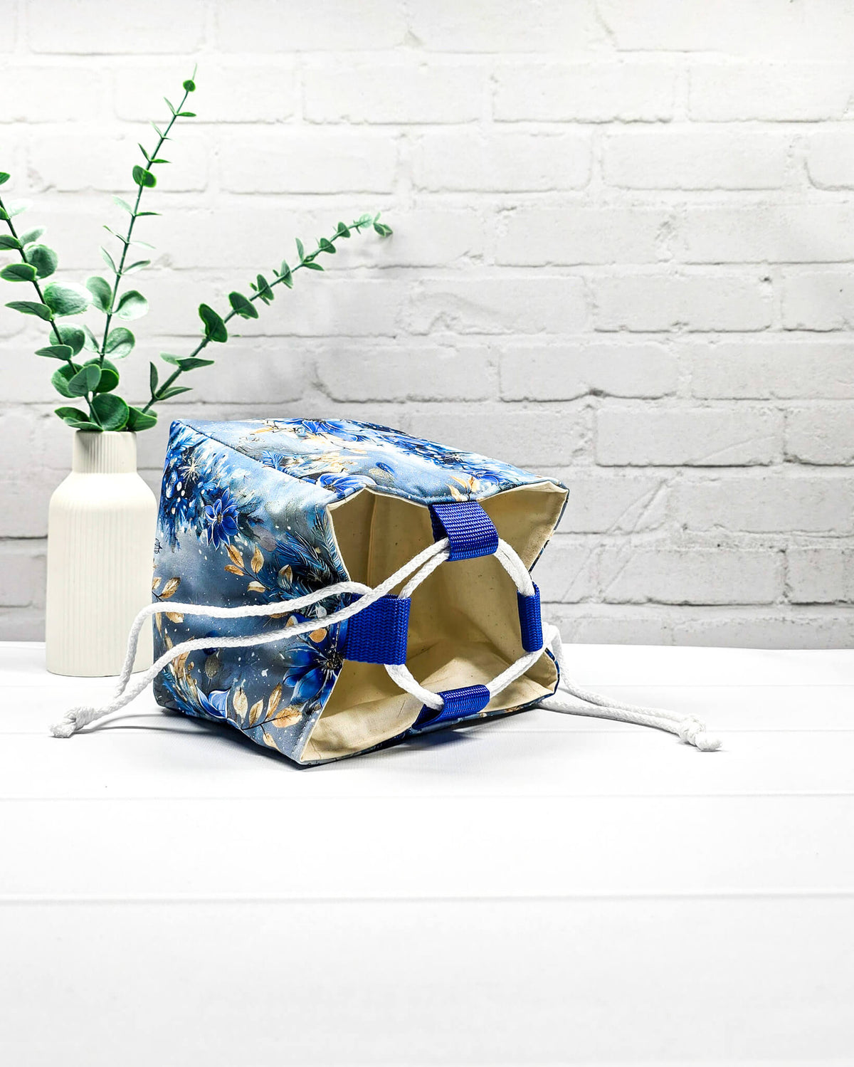 Projekttasche Aska Größe S als Rice Bag mit blauem Winterblütenmotiv und goldenen Akzenten auf hellem Hintergrund. Mit weißem Kordelzug und blauen Schlaufen. Handgenäht in Bayern von Nadelmikado.