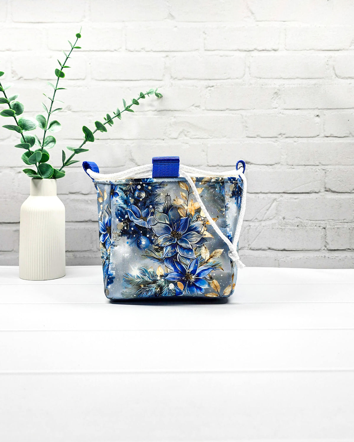 Projekttasche Aska Größe S als Rice Bag mit blauem Winterblütenmotiv und goldenen Akzenten auf hellem Hintergrund. Mit weißem Kordelzug und blauen Schlaufen. Handgenäht in Bayern von Nadelmikado.