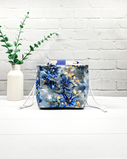 Projekttasche Aska Größe S als Rice Bag mit blauem Winterblütenmotiv und goldenen Akzenten auf hellem Hintergrund. Mit weißem Kordelzug und blauen Schlaufen. Handgenäht in Bayern von Nadelmikado.