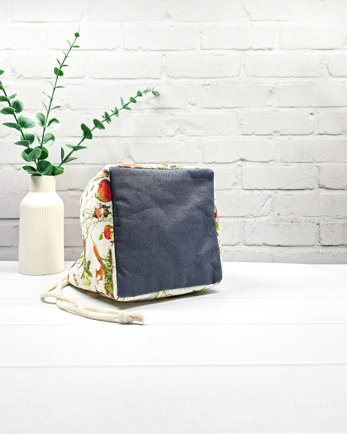 Projekttasche Aska Größe S als Rice Bag aus hellem Stoff mit Eichhörnchen, kleinen Vögeln, roten Äpfeln und feinen Waldpflanzen. Mit hellem Kordelzug und dunklem Boden. Handgenäht in Bayern von Nadelmikado.