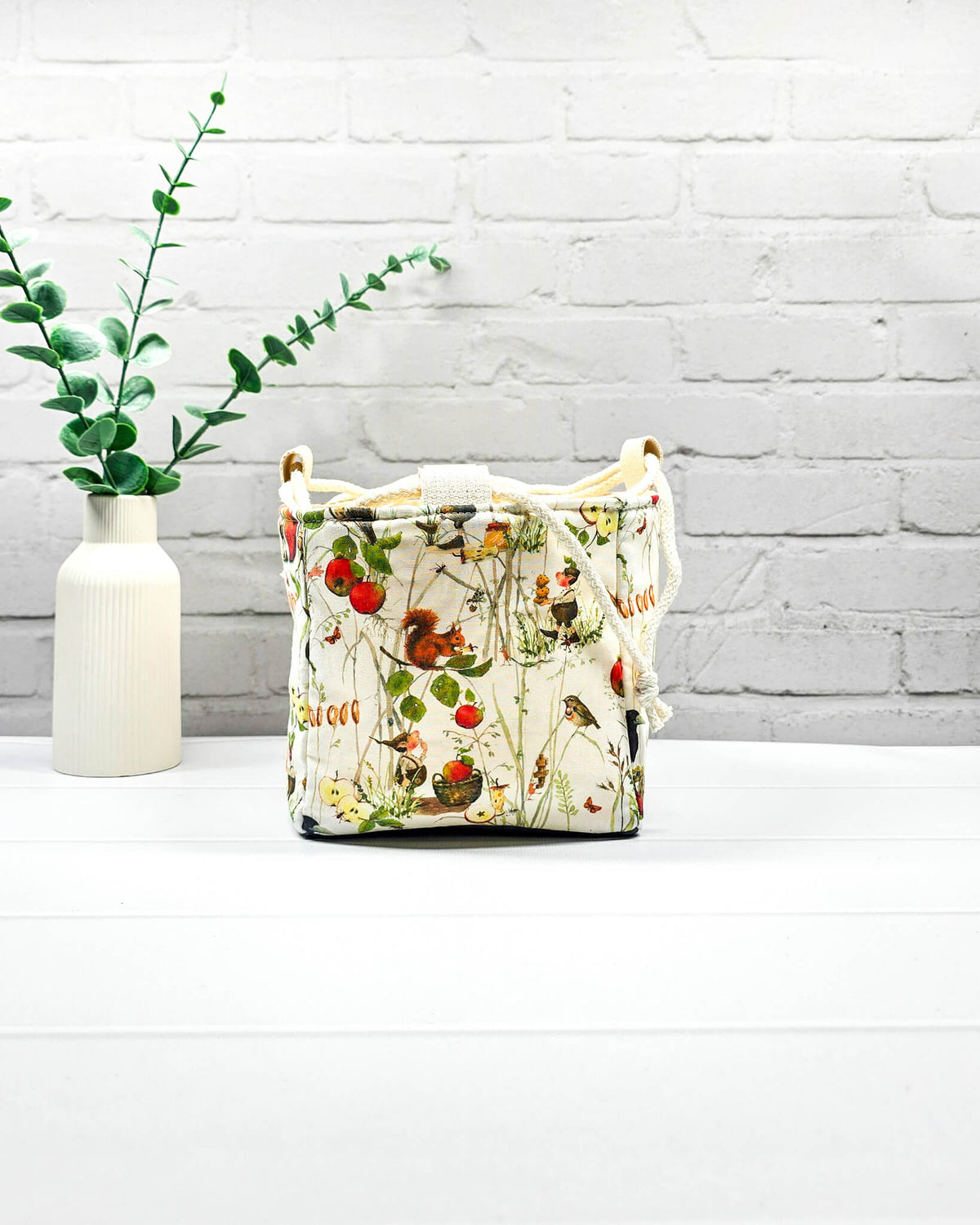 Projekttasche Aska Größe S als Rice Bag aus hellem Stoff mit Eichhörnchen, kleinen Vögeln, roten Äpfeln und feinen Waldpflanzen. Mit hellem Kordelzug und dunklem Boden. Handgenäht in Bayern von Nadelmikado.