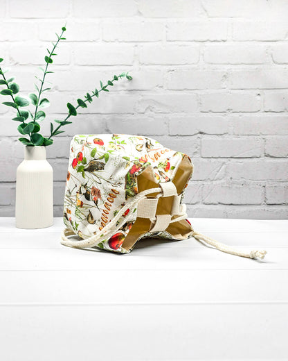 Projekttasche Aska Größe S als Rice Bag aus hellem Stoff mit Eichhörnchen, kleinen Vögeln, roten Äpfeln und feinen Waldpflanzen. Mit hellem Kordelzug und dunklem Boden. Handgenäht in Bayern von Nadelmikado.