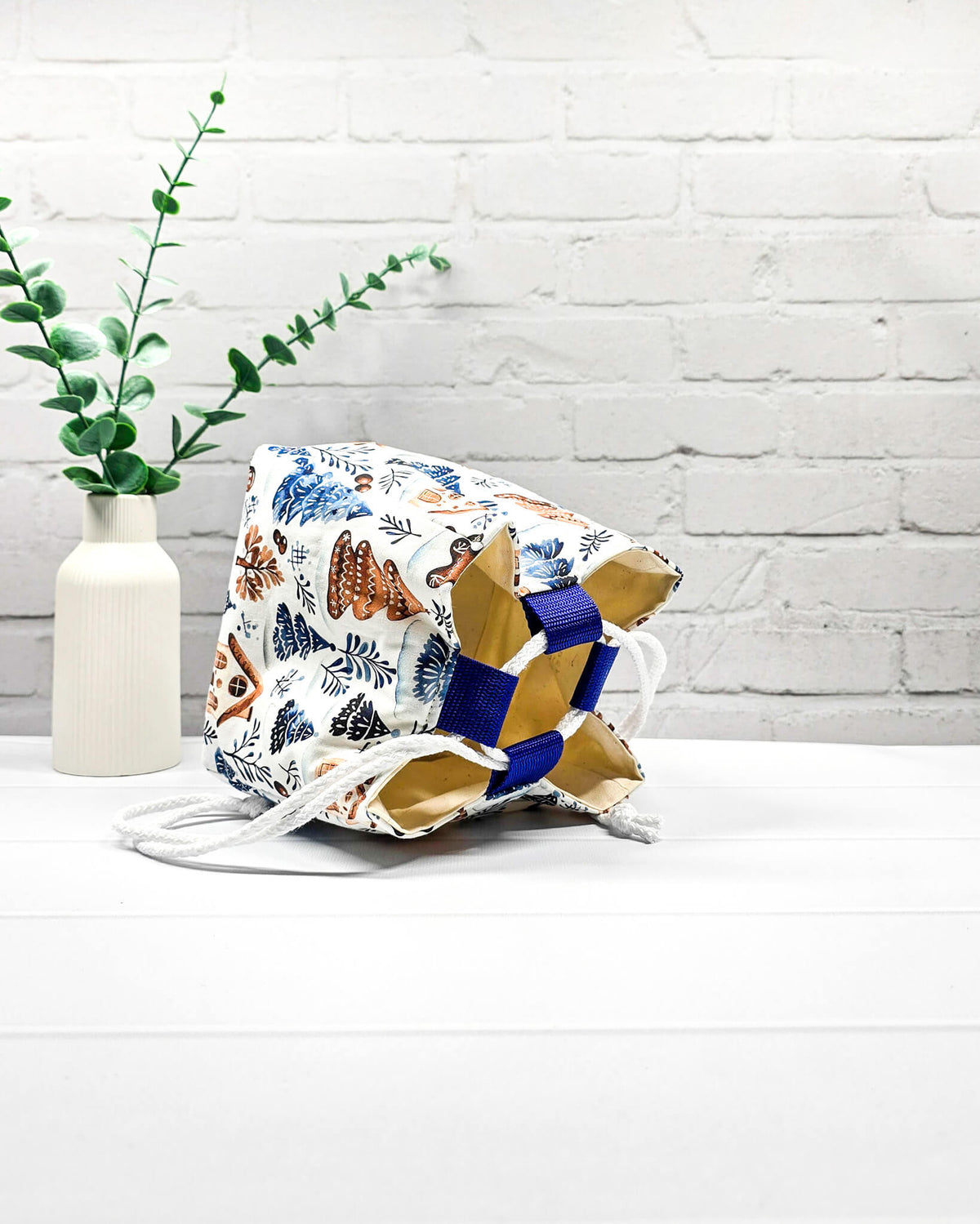 Projekttasche Aska Größe S als Rice Bag aus weißem Stoff mit blauen Tannenbäumen, Lebkuchenhäuschen und winterlichen Zweigen. Mit weißem Kordelzug und blauen Schlaufen. Handgenäht in Bayern von Nadelmikado.