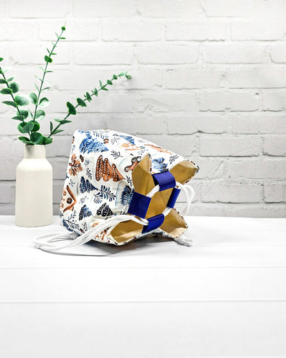 Projekttasche Aska Größe S als Rice Bag aus weißem Stoff mit blauen Tannenbäumen, Lebkuchenhäuschen und winterlichen Zweigen. Mit weißem Kordelzug und blauen Schlaufen. Handgenäht in Bayern von Nadelmikado.