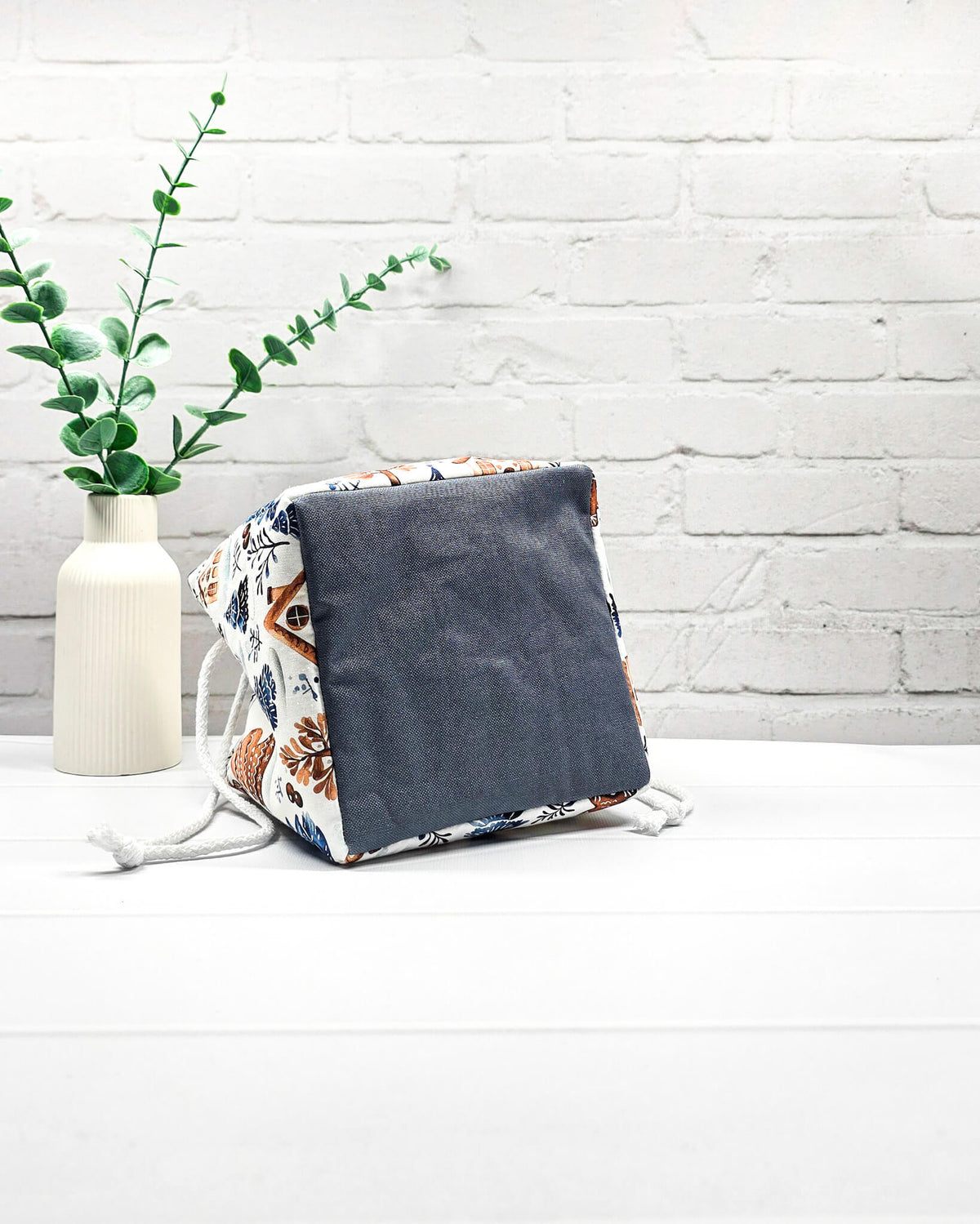 Projekttasche Aska Größe S als Rice Bag aus weißem Stoff mit blauen Tannenbäumen, Lebkuchenhäuschen und winterlichen Zweigen. Mit weißem Kordelzug und blauen Schlaufen. Handgenäht in Bayern von Nadelmikado.