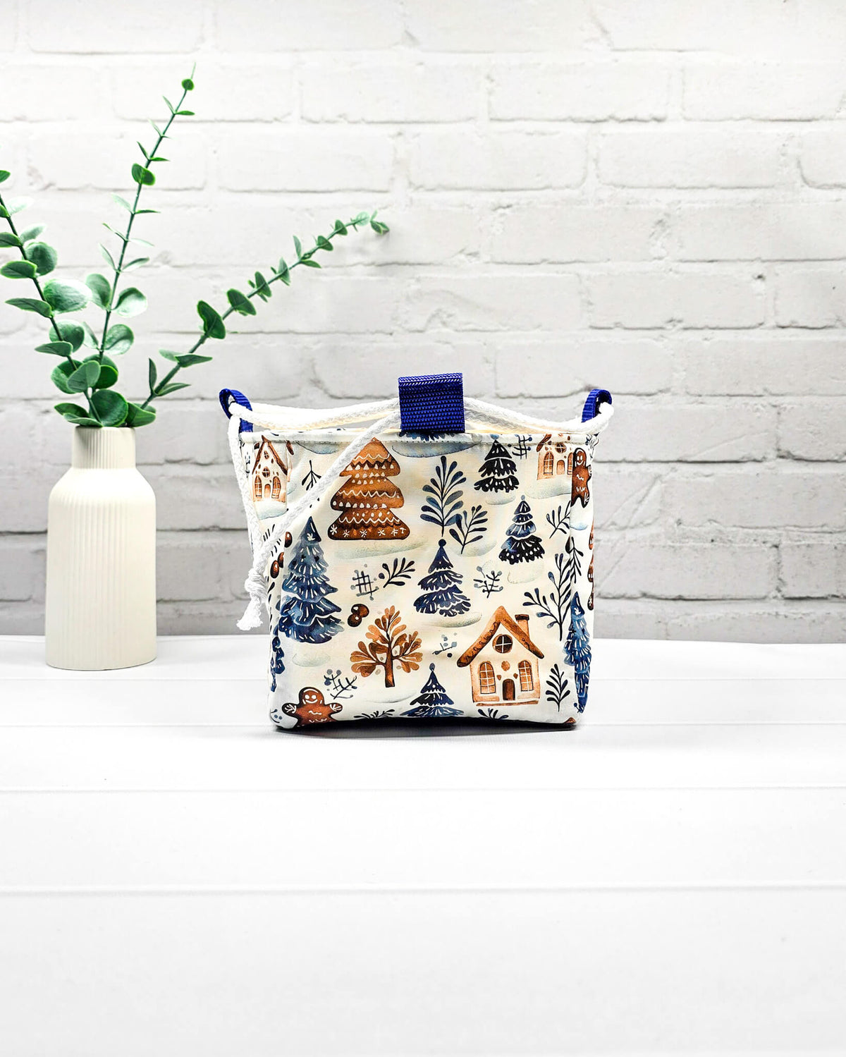 Projekttasche Aska Größe S als Rice Bag aus weißem Stoff mit blauen Tannenbäumen, Lebkuchenhäuschen und winterlichen Zweigen. Mit weißem Kordelzug und blauen Schlaufen. Handgenäht in Bayern von Nadelmikado.