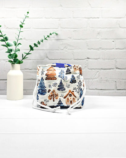 Projekttasche Aska Größe S als Rice Bag aus weißem Stoff mit blauen Tannenbäumen, Lebkuchenhäuschen und winterlichen Zweigen. Mit weißem Kordelzug und blauen Schlaufen. Handgenäht in Bayern von Nadelmikado.