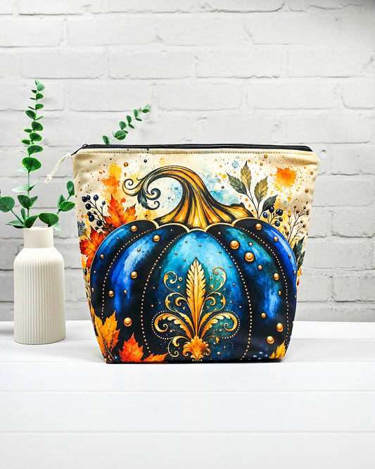 Strickbeutel Größe M mit großem blauem Kürbis, goldenen Ornamenten und Herbstlaub auf hellem Grund sowie schwarzem Reißverschluss. Handgenäht in Bayern von Nadelmikado.