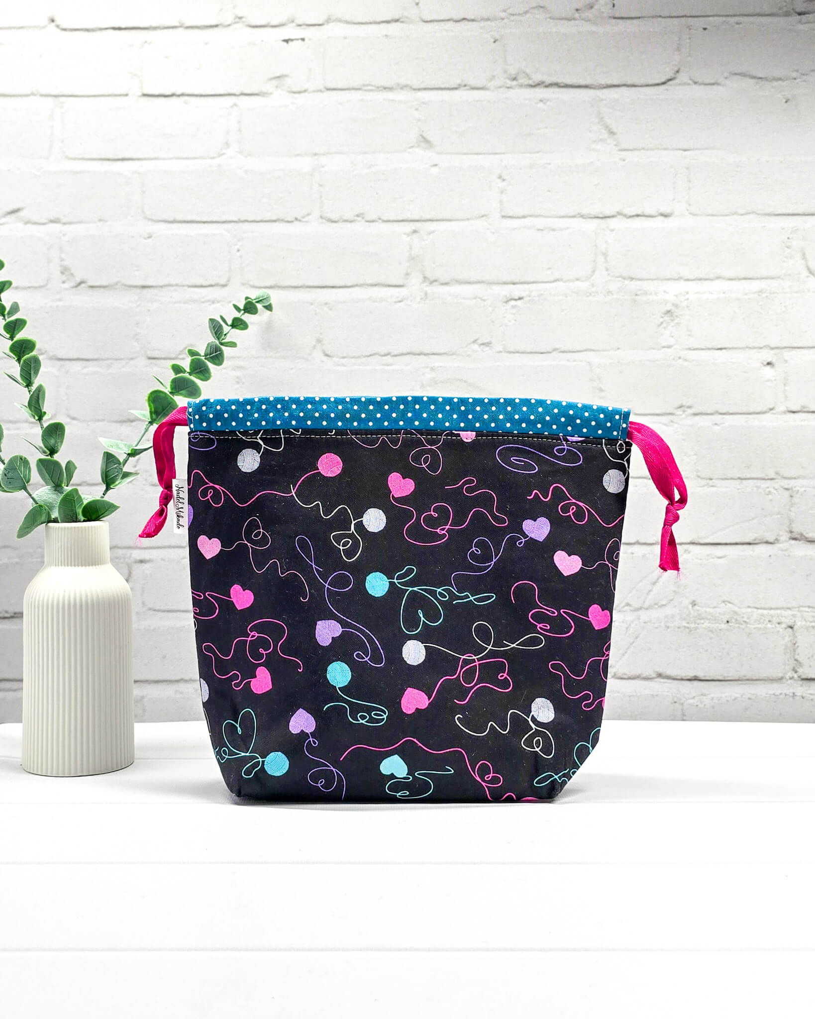 Projekttasche in Größe S aus schwarzem Stoff mit bunten Wollknäuel- und Herzmotiven, einem türkisfarbenen Pünktchen-Rand und pinken Kordelzügen. Handgenäht in Bayern von Nadelmikado. 