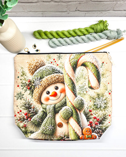Projekttasche Isvind Größe M mit fröhlichem Schneemann in grüner Strickmütze und Schal, großer grün-weißer Zuckerstange, Schneeflocken und winterlichen Tannenzweigen. Mit Reißverschluss für Strickprojekte bis 400 g Wolle. Handgenäht in Bayern von Nadelmikado.
