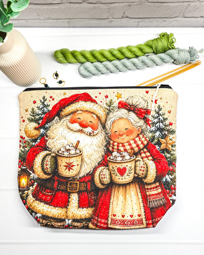 Projekttasche Isvind Größe M mit fröhlichem Weihnachtsmann und Weihnachtsfrau in rot-weißer Winterkleidung, heißen Kakaotassen, Tannenbäumen und Weihnachtssternen. Mit Reißverschluss für Strickprojekte bis 400 g Wolle. Handgenäht in Bayern von Nadelmikado.