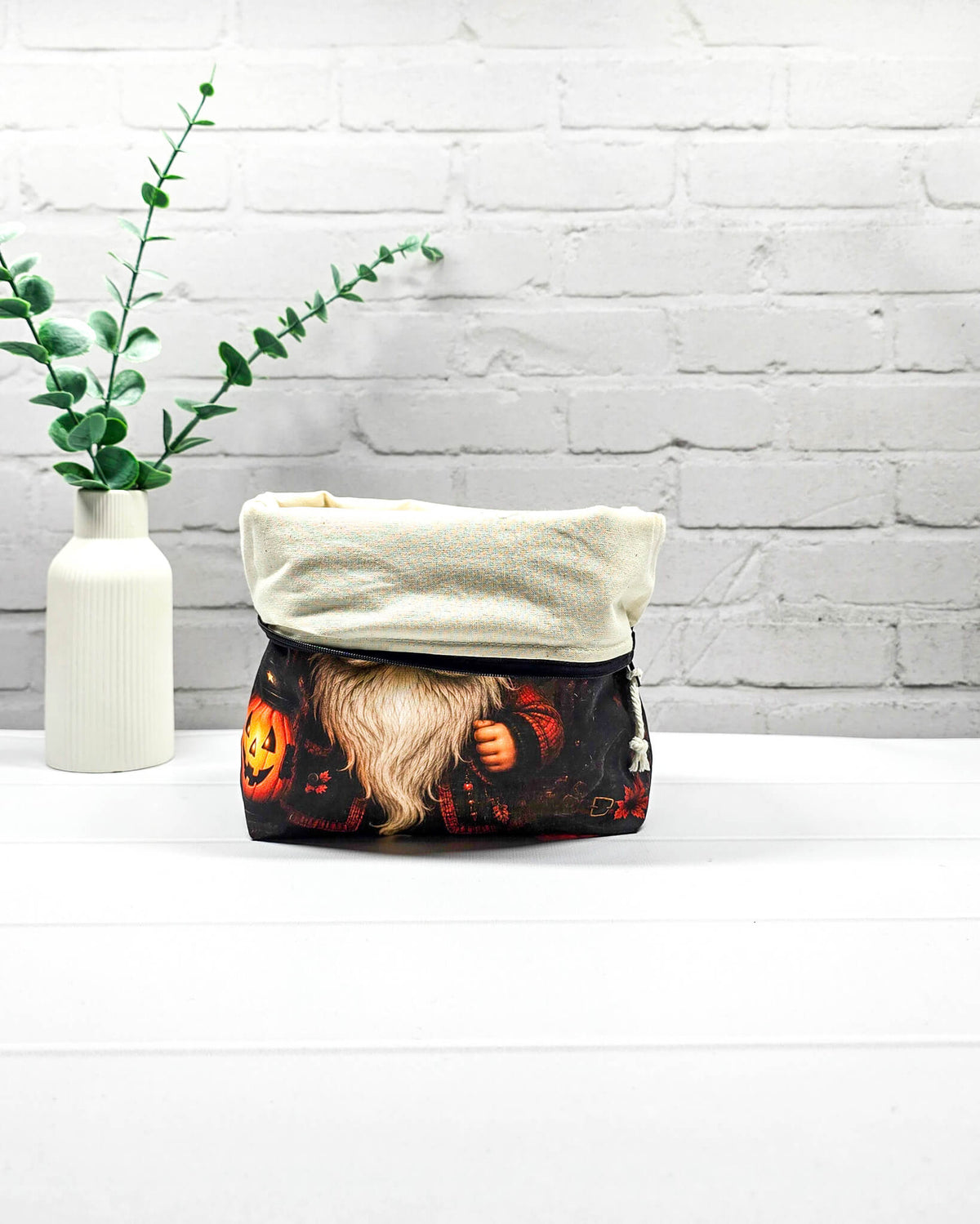 Projekttasche Isvind Größe S mit Halloweenwichtel, schwarzer Zipfelmütze, weißem Bart und leuchtender Kürbislaterne auf dunklem Hintergrund. Mit Reißverschluss für kleine Strickprojekte. Handgenäht in Bayern von Nadelmikado.