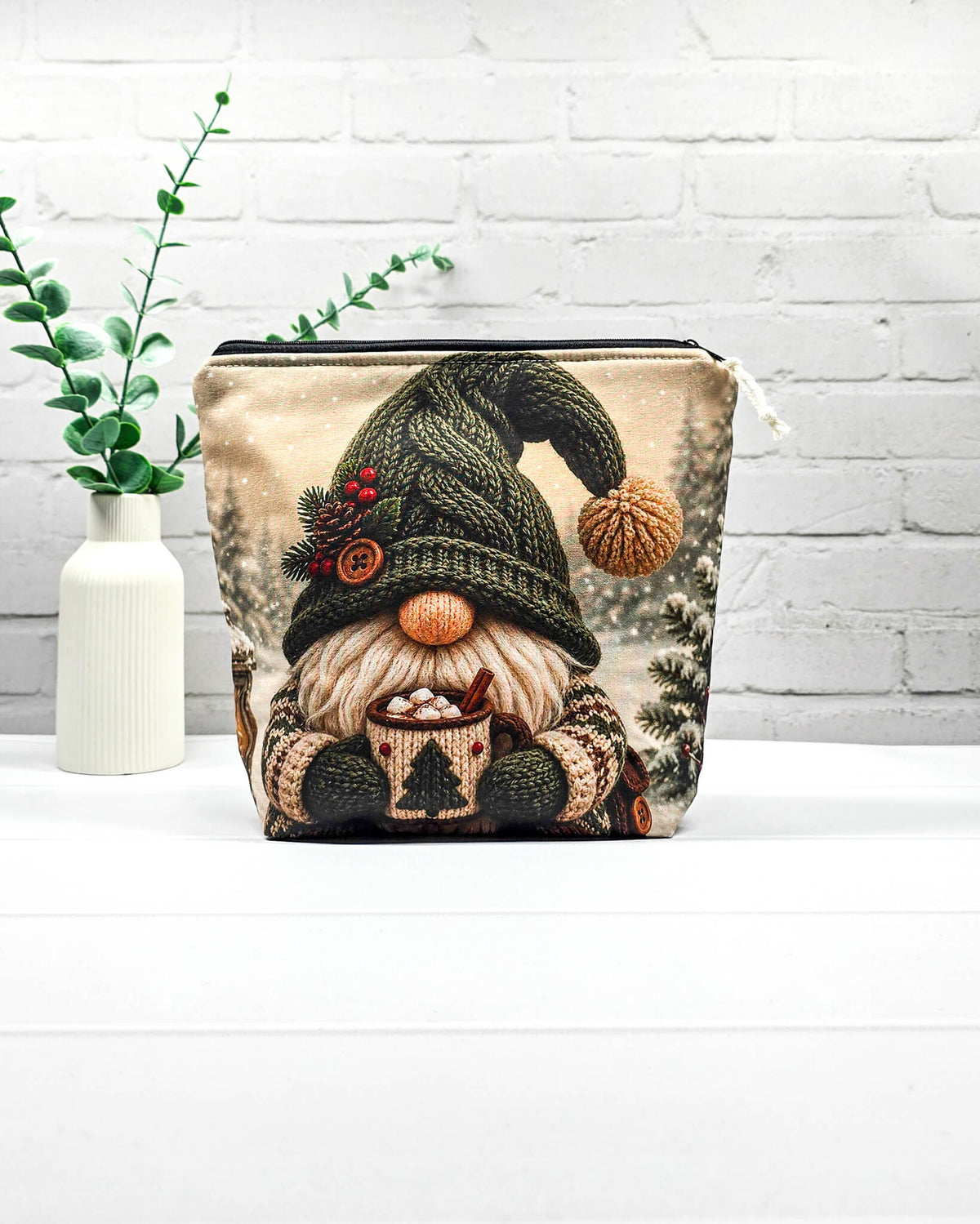 Projekttasche Isvind Größe S mit Winterwichtel, großer grüner Strickmütze, weißem Bart und einer Tasse heißem Kakao vor verschneiten Tannenbäumen. Mit Reißverschluss für kleine Strickprojekte. Handgenäht in Bayern von Nadelmikado.