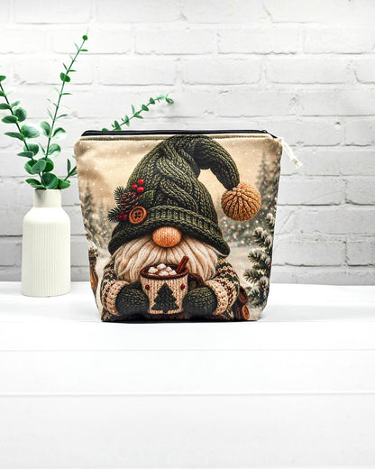 Projekttasche Isvind Größe S mit Winterwichtel, großer grüner Strickmütze, weißem Bart und einer Tasse heißem Kakao vor verschneiten Tannenbäumen. Mit Reißverschluss für kleine Strickprojekte. Handgenäht in Bayern von Nadelmikado.