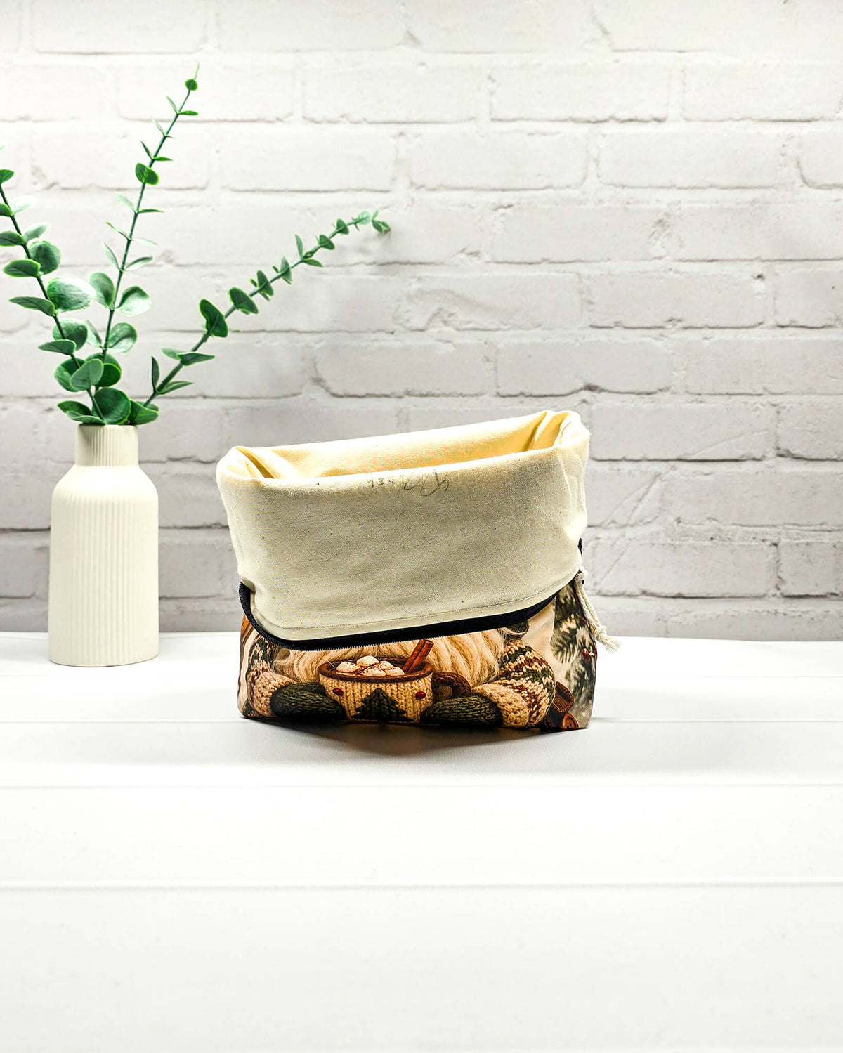Projekttasche Isvind Größe S mit Winterwichtel, großer grüner Strickmütze, weißem Bart und einer Tasse heißem Kakao vor verschneiten Tannenbäumen. Mit Reißverschluss für kleine Strickprojekte. Handgenäht in Bayern von Nadelmikado.