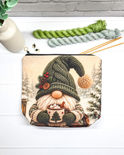 Projekttasche Isvind Größe S mit Winterwichtel, großer grüner Strickmütze, weißem Bart und einer Tasse heißem Kakao vor verschneiten Tannenbäumen. Mit Reißverschluss für kleine Strickprojekte. Handgenäht in Bayern von Nadelmikado.