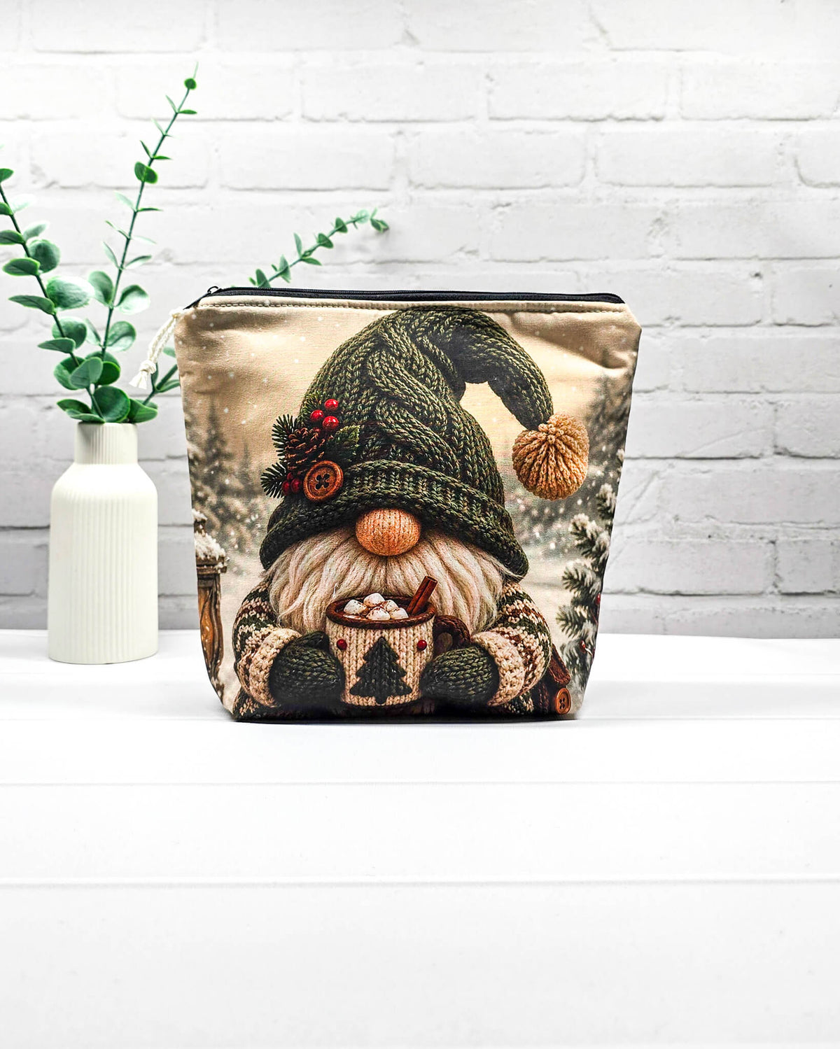 Projekttasche Isvind Größe S mit Winterwichtel, großer grüner Strickmütze, weißem Bart und einer Tasse heißem Kakao vor verschneiten Tannenbäumen. Mit Reißverschluss für kleine Strickprojekte. Handgenäht in Bayern von Nadelmikado.