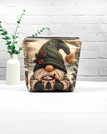 Projekttasche Isvind Größe S mit Winterwichtel, großer grüner Strickmütze, weißem Bart und einer Tasse heißem Kakao vor verschneiten Tannenbäumen. Mit Reißverschluss für kleine Strickprojekte. Handgenäht in Bayern von Nadelmikado.